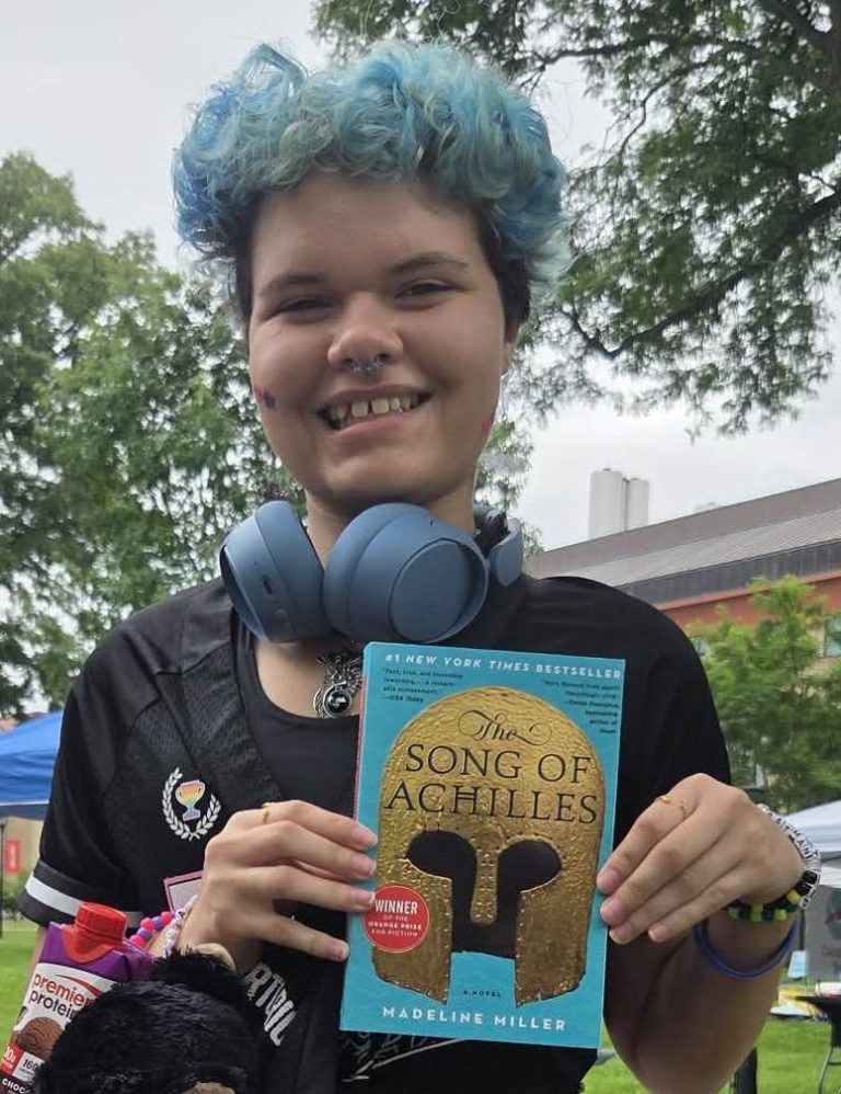 Young person with blue hair and headphones around their neck holds the book The Song of Achilles